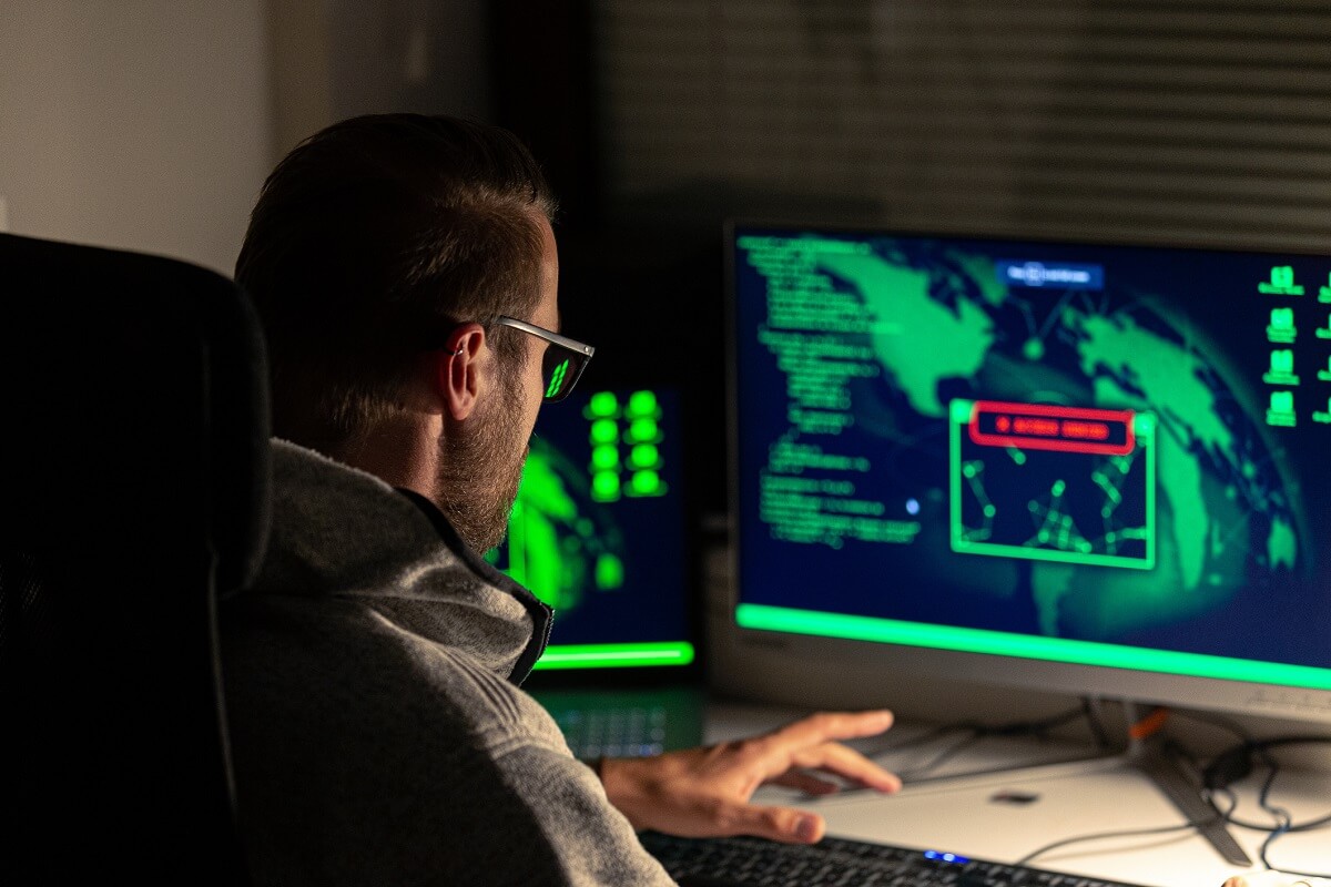 A man in a dimly-lit room stares into the monitor on his desk. The computer screen has a map and green code and a red warning.