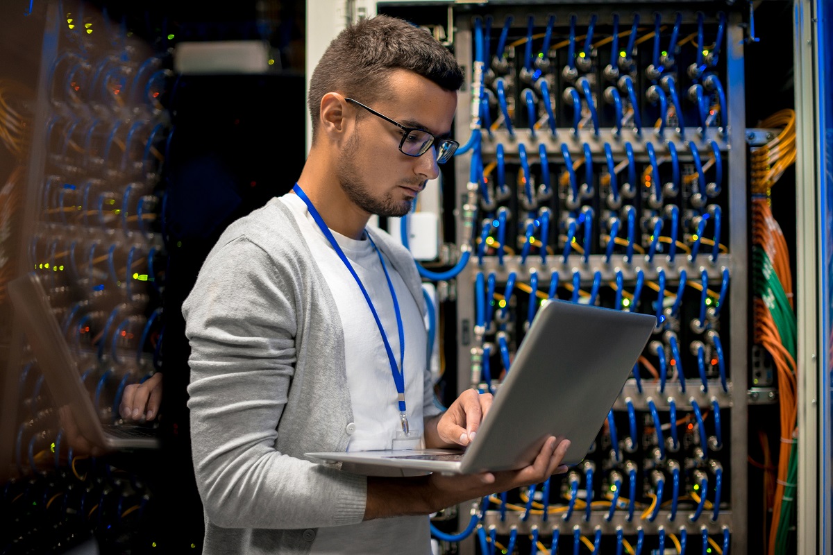 Young man with laptop standing by server cabinet while working with supercomputer