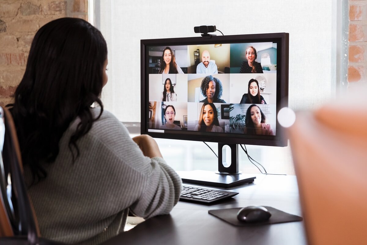 Back of a businesswoman in her home office meets with colleagues on a web conference, with a grid of faces on her computer. 