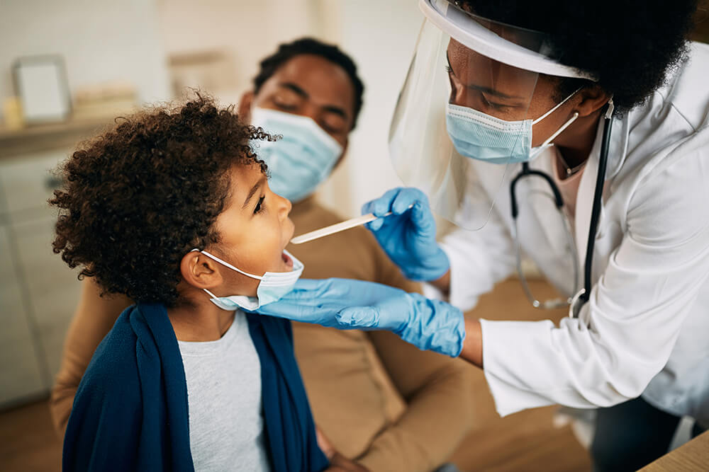 African American nurse practitioner with face mask examining small African American boy\'s throat during a home visit.